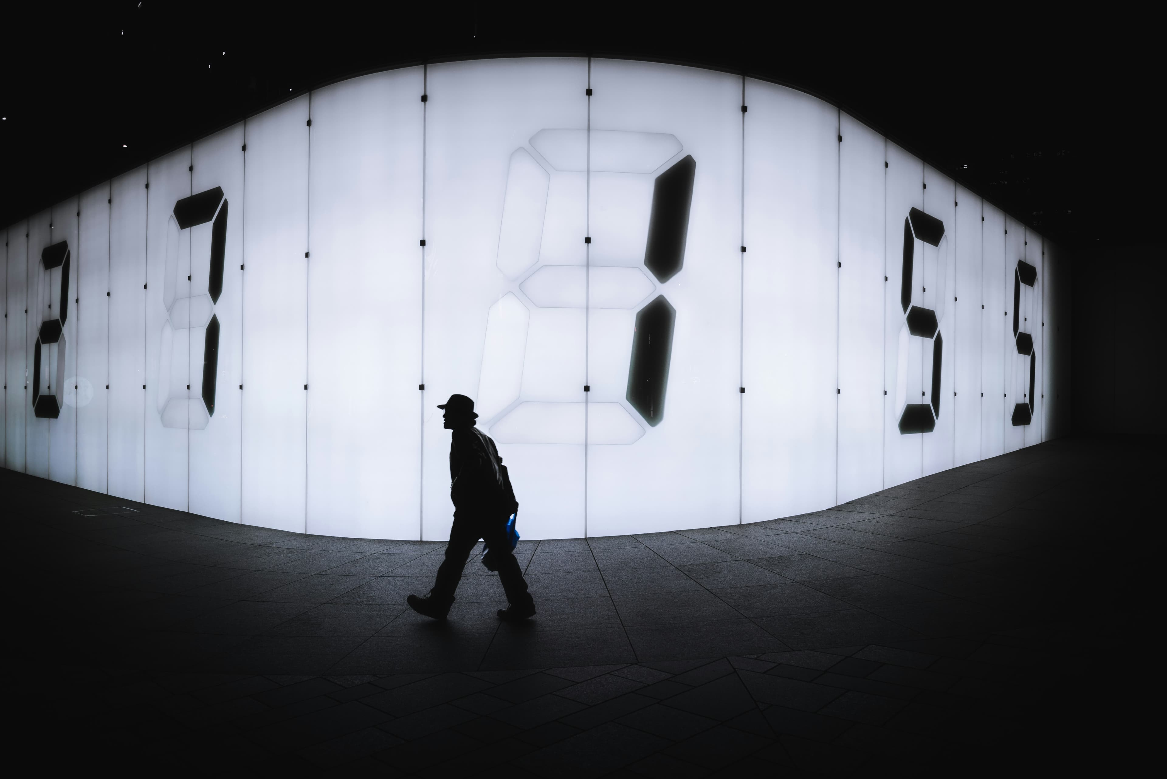 A person walking past a wall of clocks, suggesting time and long-term decisions